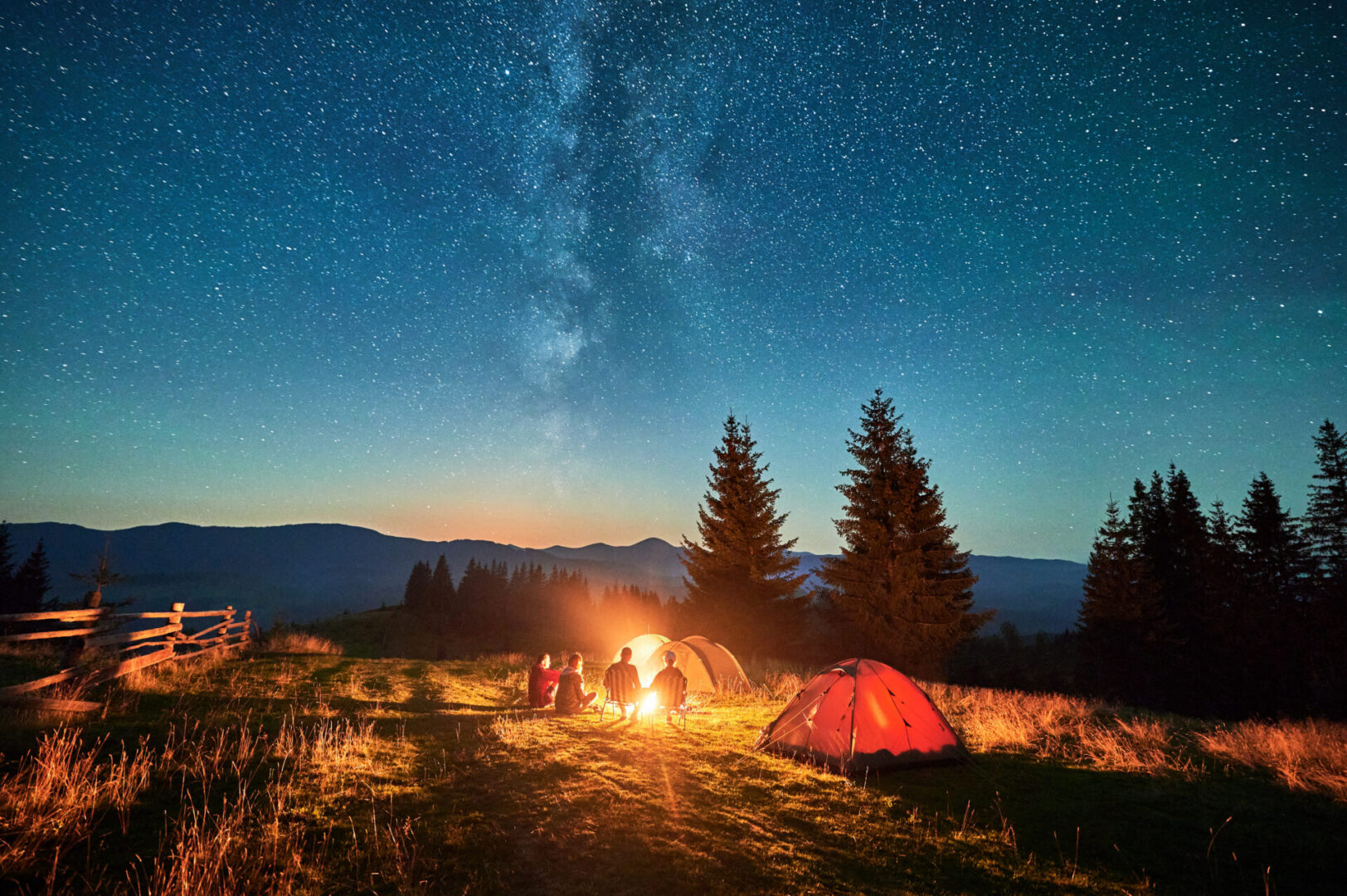 Group of friends hikers resting around campfire, surrounded by illuminated tents under stunning starry sky. Milky Way stretches above, creating magical atmosphere in peaceful mountain wilderness.
